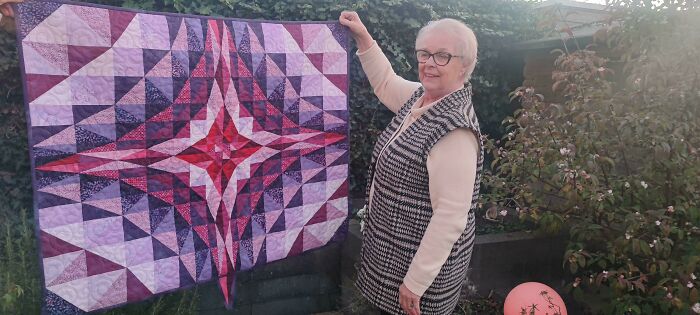 Elderly woman proudly displaying a colorful, geometric quilt outdoors, symbolizing amazing mothers-in-law.