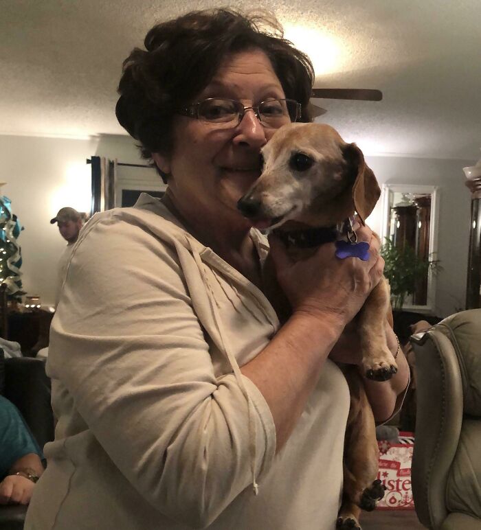 A smiling woman, a loving mother-in-law, holding a brown dog in a cozy living room setting.