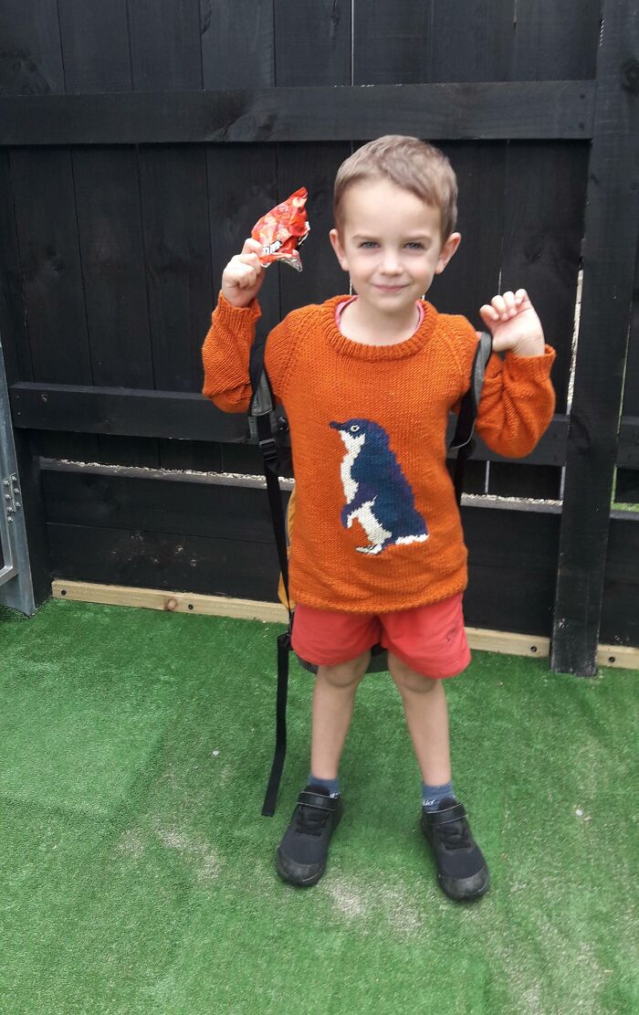 Child in an orange penguin sweater holding a snack, standing on a green grassy area, smiling.