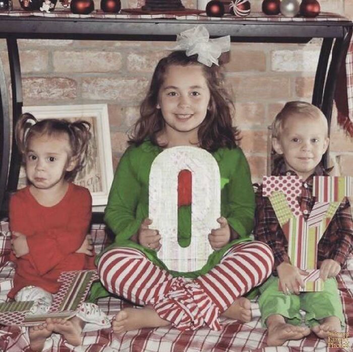 Three children sitting, holding letters spelling "JOY," dressed in festive attire, awkward family photo setting.