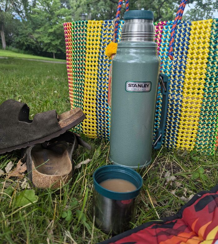 Colorful woven bag, green Stanley thermos with cup of coffee, and worn sandals on grass, showing a cool inheritance item.