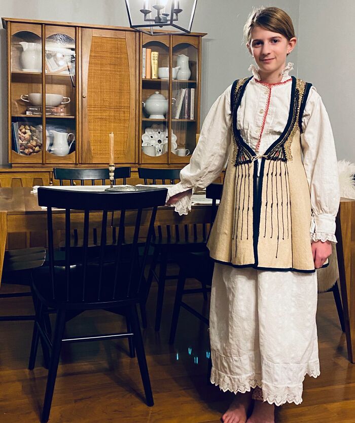 Young person in traditional vintage clothing standing next to antique wooden dining table and chairs, showcasing cool inheritance.