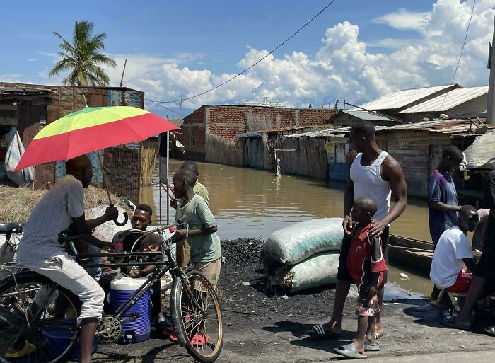 Urban hell scene with a group of people near flooded streets, surrounded by makeshift homes under a colorful umbrella.