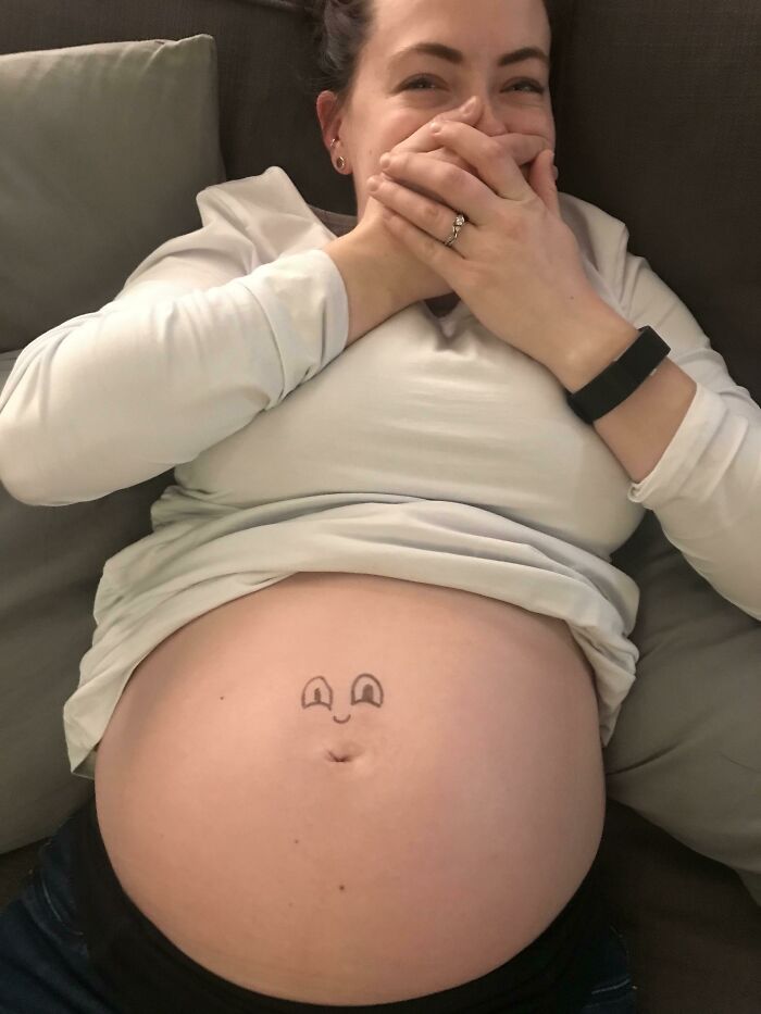 Pregnant belly with smiley face drawing, woman laughing with hands over mouth.