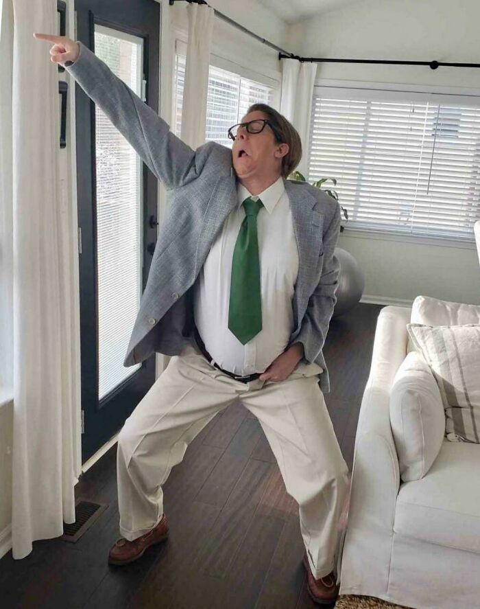 Person in a suit humorously posed, mimicking a pregnancy belly, with an exaggerated expression in a living room.