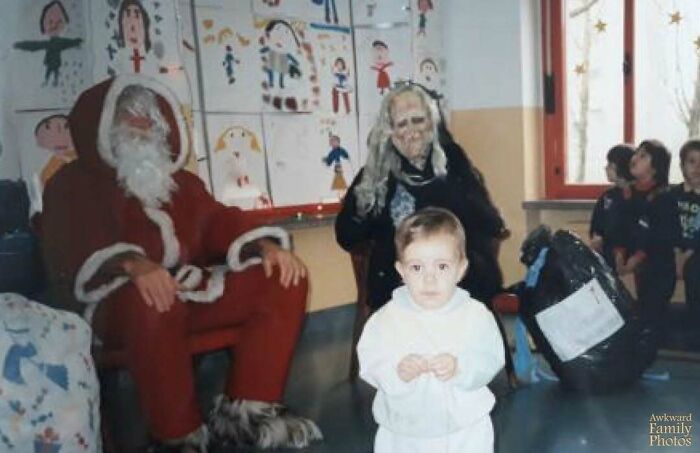 Awkward family photo with a child, Santa, and a person in a spooky costume, in a classroom setting with children’s drawings.
