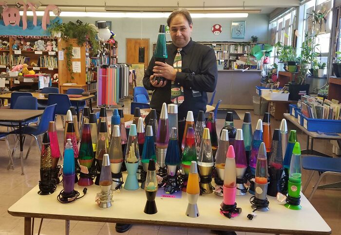 A man stands behind a table displaying a collection of colorful lava lamps in a cluttered room.