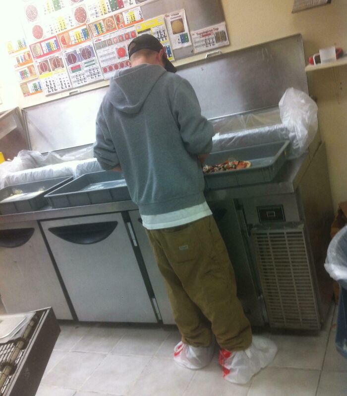Employee in a kitchen wearing shoe covers, preparing food with a humorous sign of good-funny-bosses on the wall.