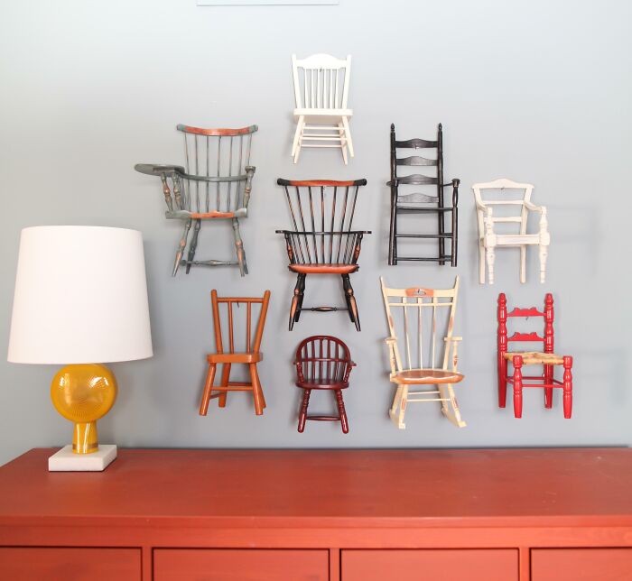 Unique collections of miniature chairs mounted on a wall above a red dresser with a lamp.