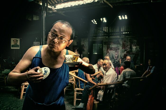 Man in dimly lit tea house, holding a cup, surrounded by seated patrons; captivating photograph by Hamed AlGhanboosi.