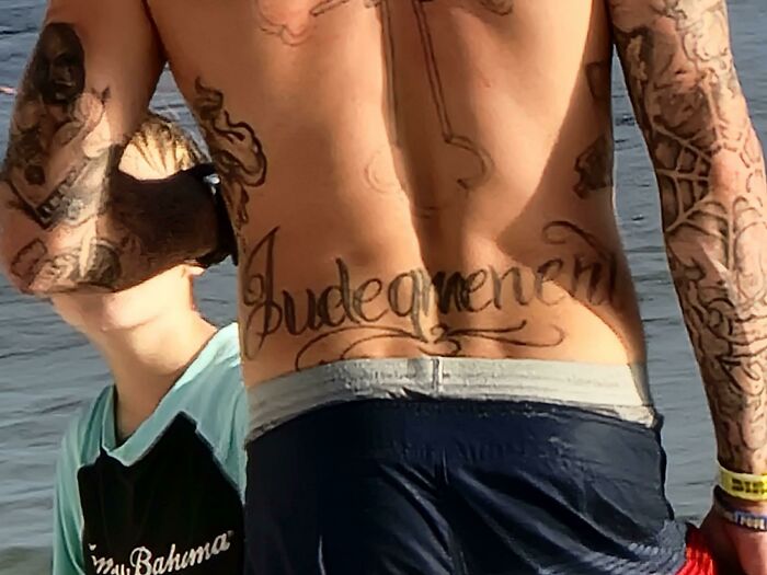 Man with "Judgement" tattoo on back, swimming at the beach; showcases funny worst tattoo fails.