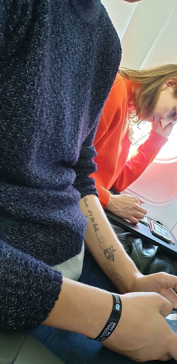 Person sitting with a funny tattoo fail on their arm, reading "never it's too late," next to another person using a phone.