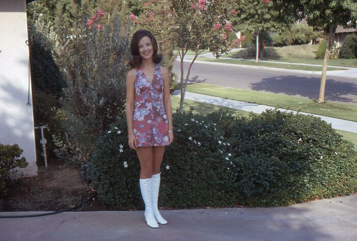 Woman in a floral mini dress and white boots, showcasing 60s fashion looks, standing in a garden setting.