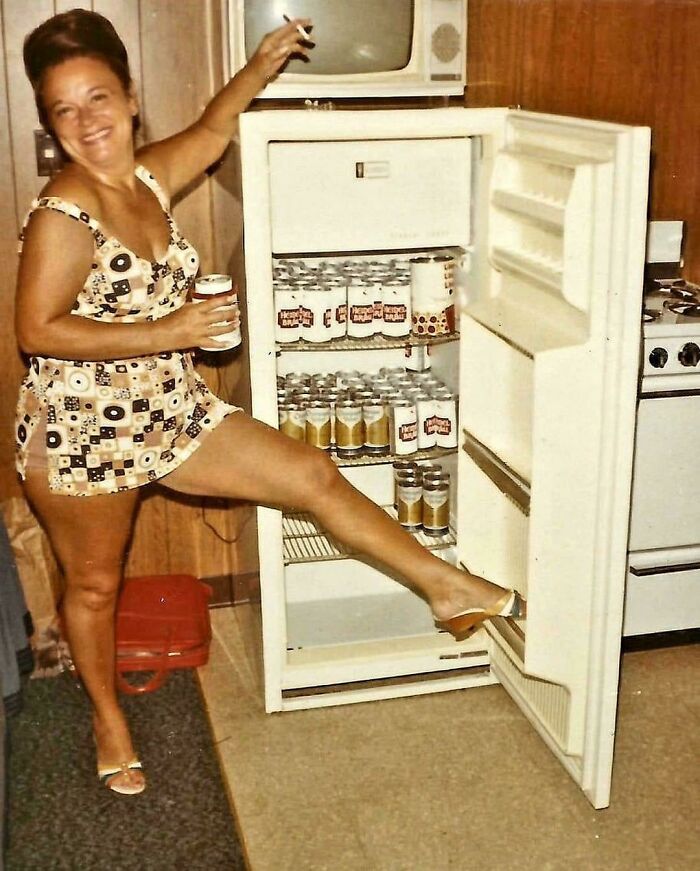 Woman in retro 60s fashion holding a drink and posing playfully with an open refrigerator.