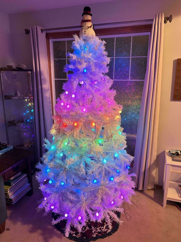 White Christmas tree with colorful lights, topped with a snowman, in a cozy living room setting.