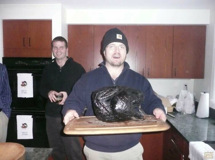Man holding a charred turkey in a kitchen, smiling awkwardly, with another man in the background. Awkward family photo.