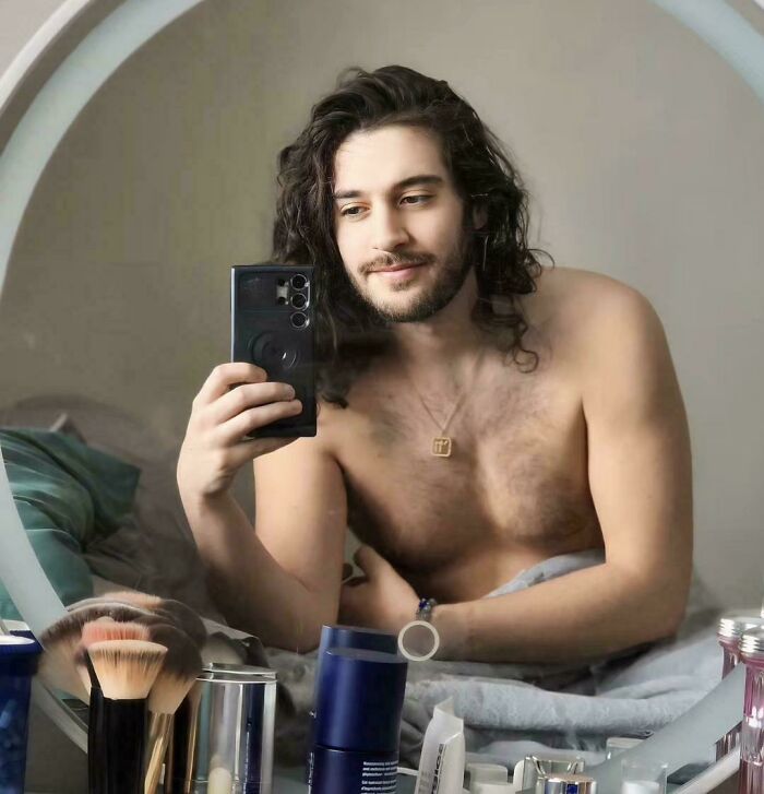 Man with fierceflow beautiful hair taking a shirtless mirror selfie in a bedroom with grooming products on the table.