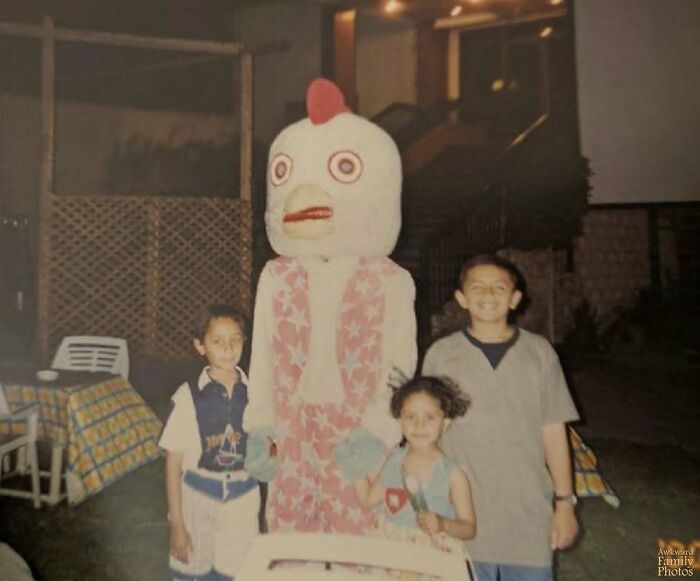 Awkward family photo with children posing next to a person in a chicken costume at an outdoor evening event.