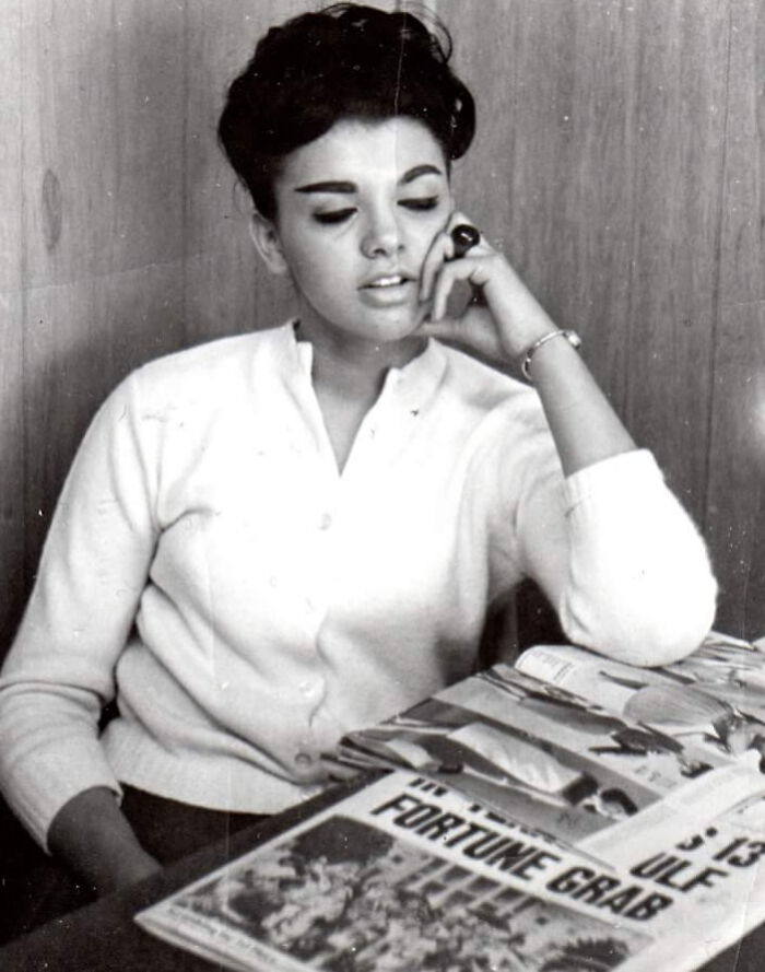 Woman in 60s fashion look, wearing a white cardigan, sitting by magazines, with an elegant hairstyle and jewelry.