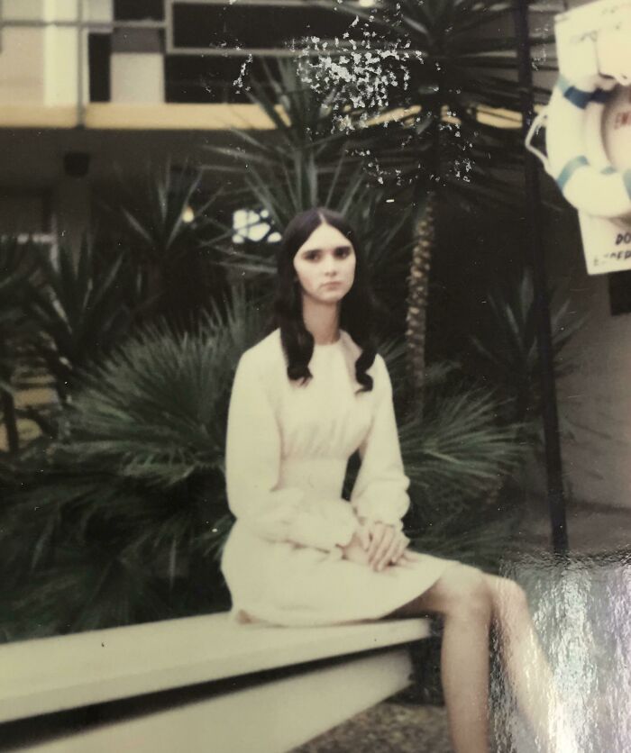 A woman in 60s fashion, wearing a vintage white dress, sitting on a bench with lush greenery in the background.
