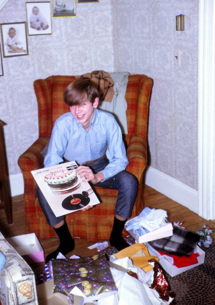 Teen playing a vinyl record, sitting in a vintage plaid chair, surrounded by gifts, showcasing 60s fashion looks.