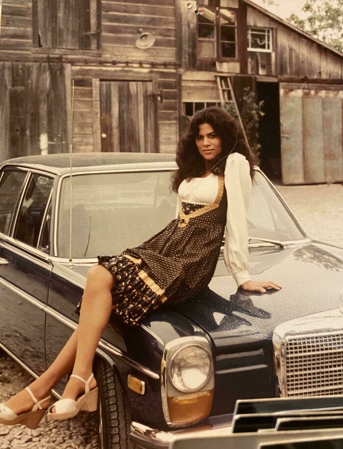 Woman in 60s fashion sitting on a vintage car, wearing a patterned dress and platform heels, with a rustic barn in the background.