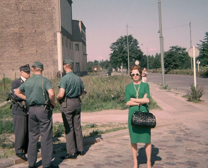 Woman in 60s fashion, wearing a green dress and sunglasses, standing on a sidewalk near three policemen.