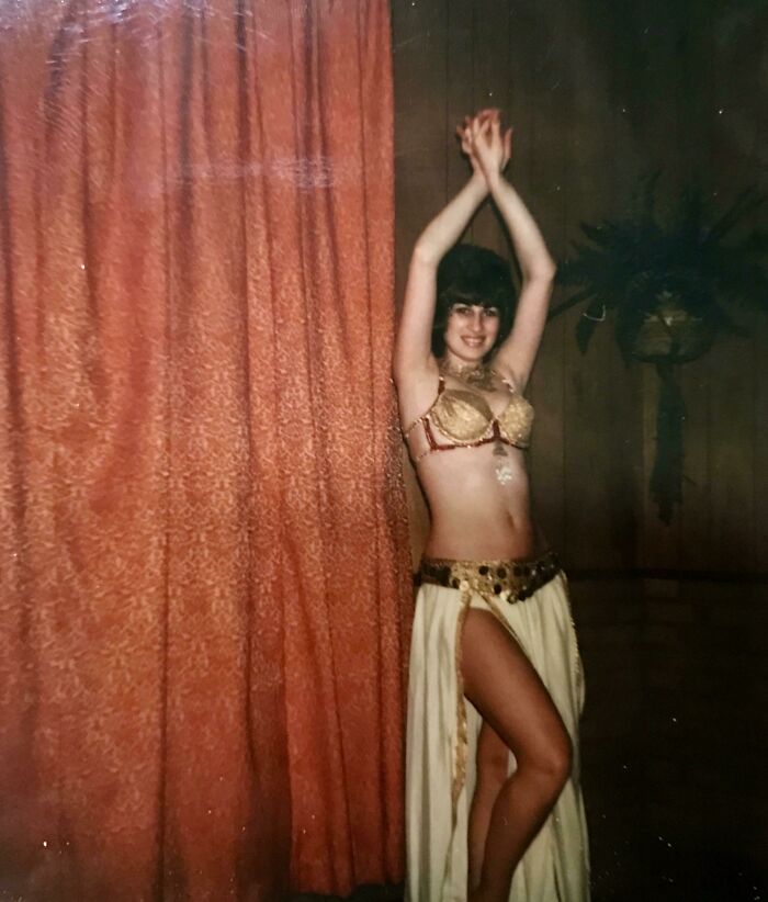 Woman showcasing 60s fashion looks in a glamorous gold and white costume, standing by an orange curtain.