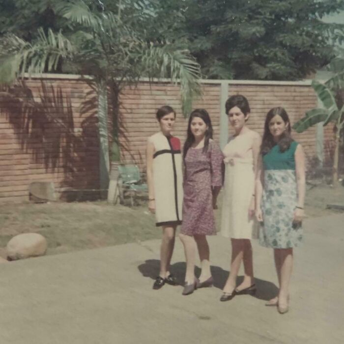 Four women showcasing 60s fashion looks in a garden, wearing vintage dresses with a brick wall and palm trees in the background.