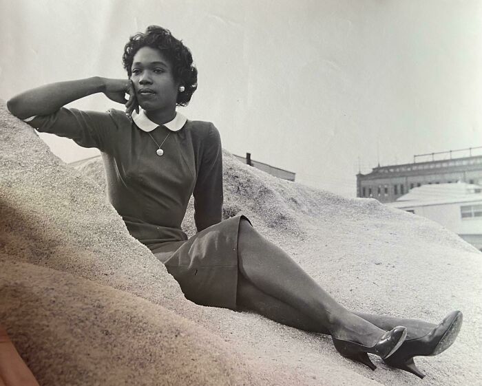 A woman in elegant 60s fashion, wearing a dress with a white collar, seated on a sandy dune.