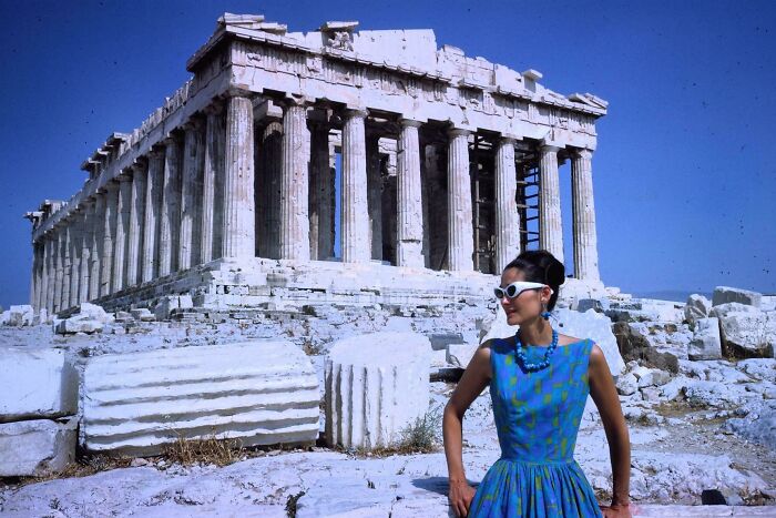 Woman in 60s fashion, wearing a blue dress and sunglasses, stands near ancient ruins under a clear blue sky.