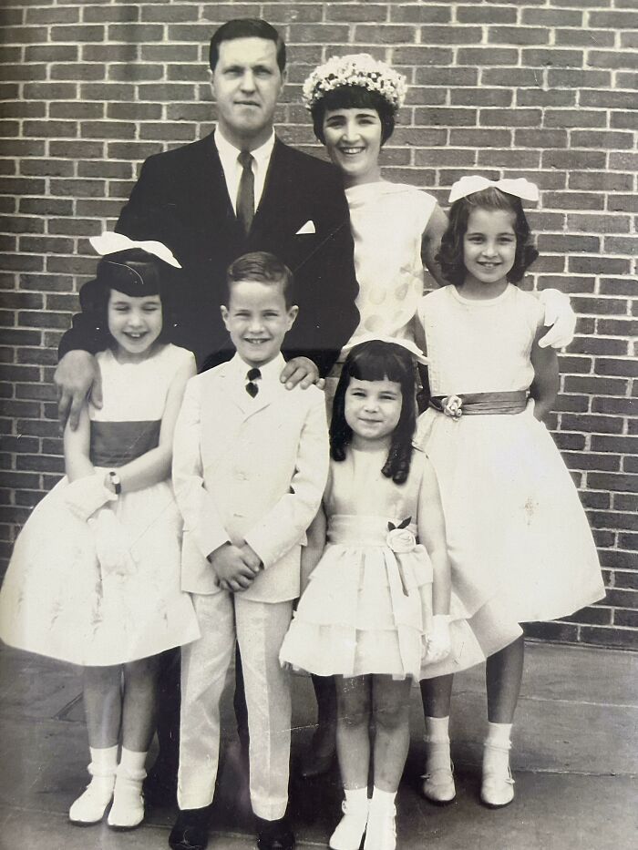 Family in 60s fashion with vintage dresses and suits, standing against a brick wall.