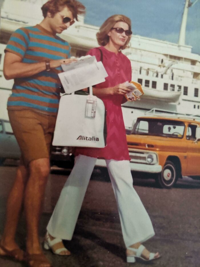 Couple in 60s fashion walking with Alitalia bag, near vintage cars and a ship in the background.