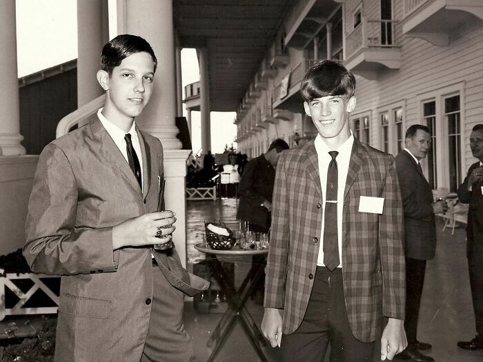 Young men in 60s fashion looks, wearing suits and ties, standing at an outdoor event, with tables in the background.