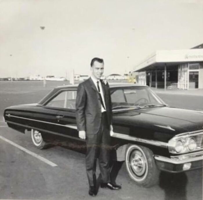 Man in a 60s fashion suit standing beside a vintage car in a parking lot.