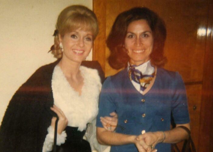 Two women in stylish 60s fashion, one with a fur-trimmed coat, the other in a blue dress with a scarf.