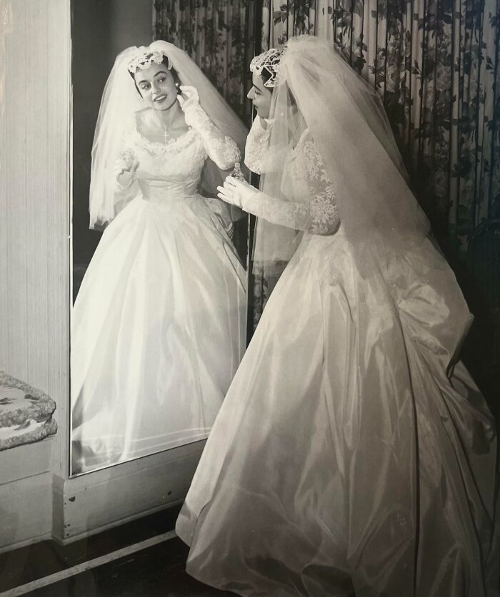 Bride in 60s fashion, wearing a vintage wedding gown with lace and veil, smiling while adjusting earrings in front of a mirror.