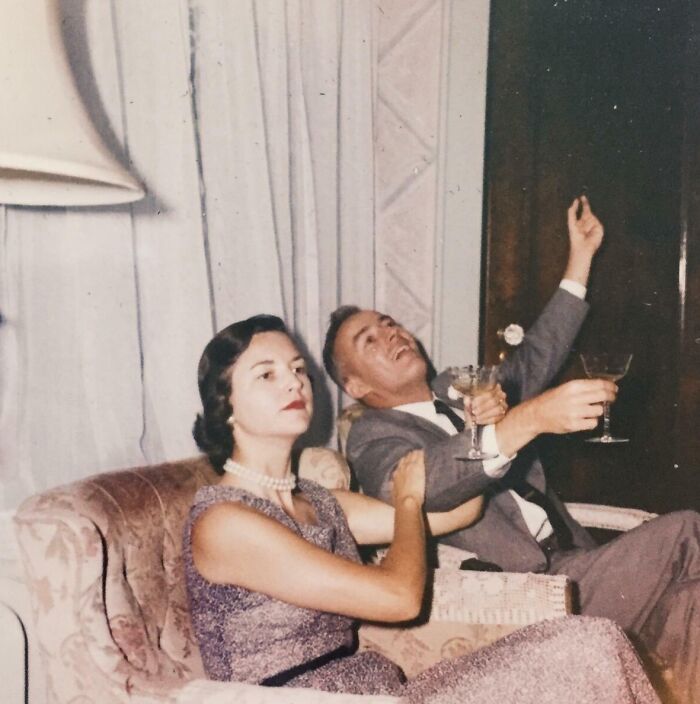 Vintage 60s fashion look with a woman in a pearl necklace and a man in a suit enjoying cocktails on a sofa.