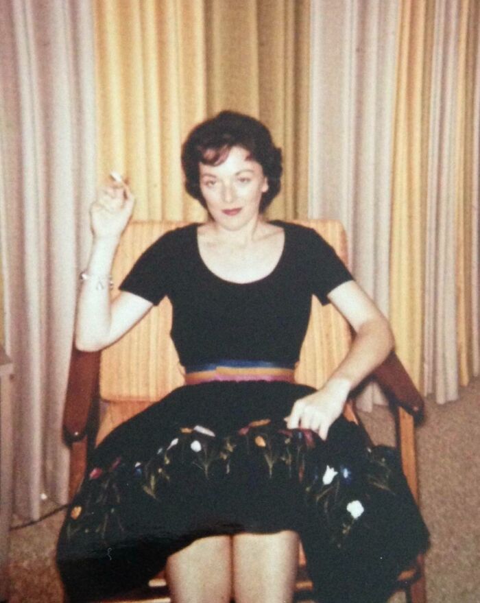 Woman in 60s fashion with a black top and embroidered skirt, seated in a vintage chair.