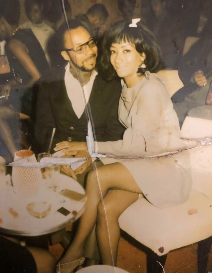 A couple in 60s fashion looks, with the woman in a stylish dress and bow, sitting at a table in a lively setting.