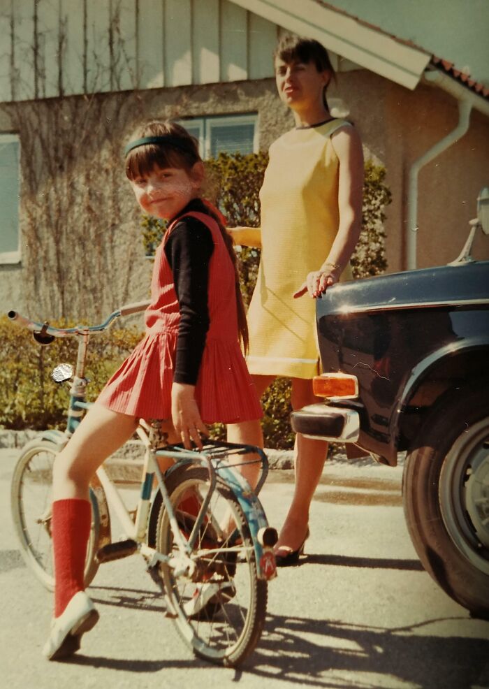 Child on a bike and woman in a yellow dress showcasing 60s fashion looks outside beside a car.