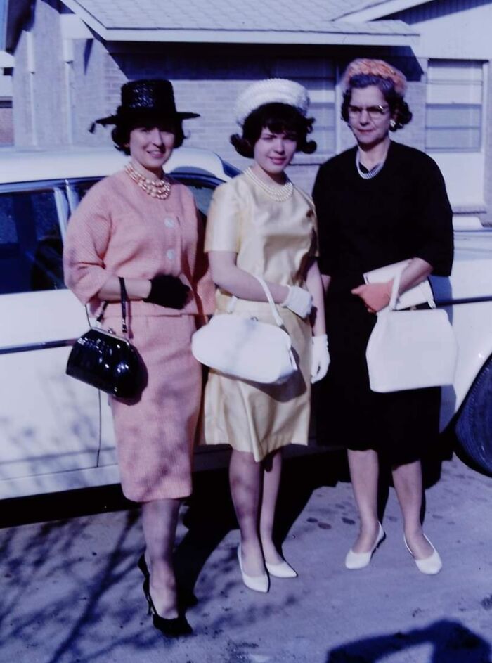 Three women in vintage 60s fashion, wearing stylish dresses, hats, and gloves, stand in front of a classic car.