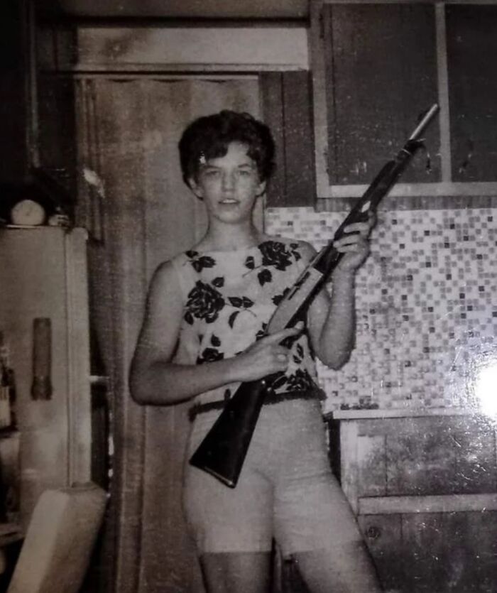 Person in 60s fashion wearing a floral top and shorts, holding a rifle in a vintage kitchen setting.