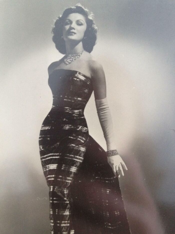 A woman in a glamorous 60s fashion dress, wearing elegant gloves and jewelry, poses confidently.