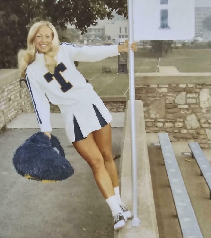 Cheerleader in 60s fashion look, wearing a vintage uniform with pom-poms outside on bleachers.
