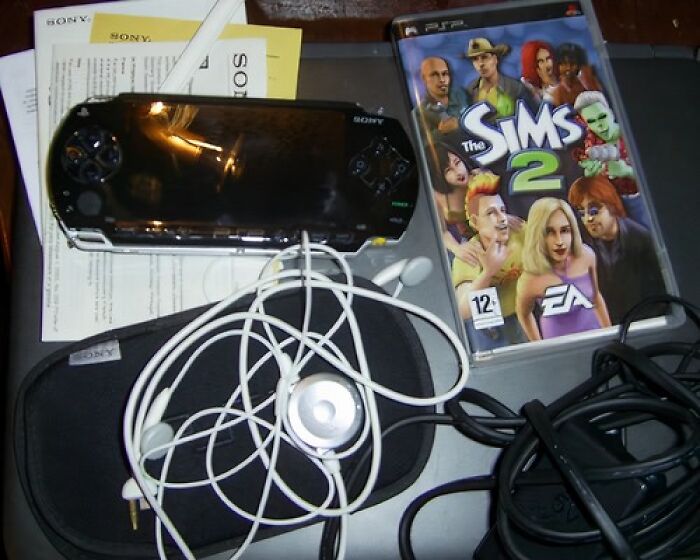 Old internet feel pic with a Sony PSP, tangled headphones, and The Sims 2 game cover on a table.