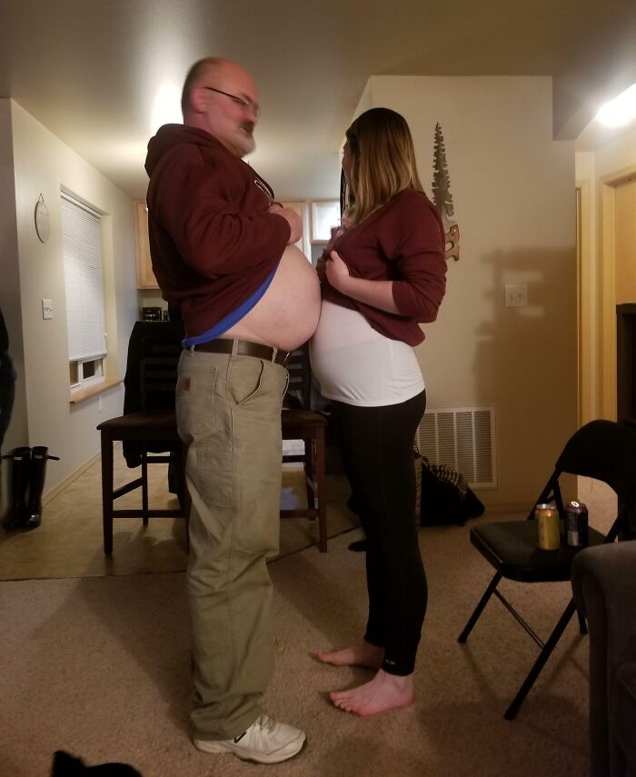 Man and pregnant woman humorously comparing bellies in a living room setting.