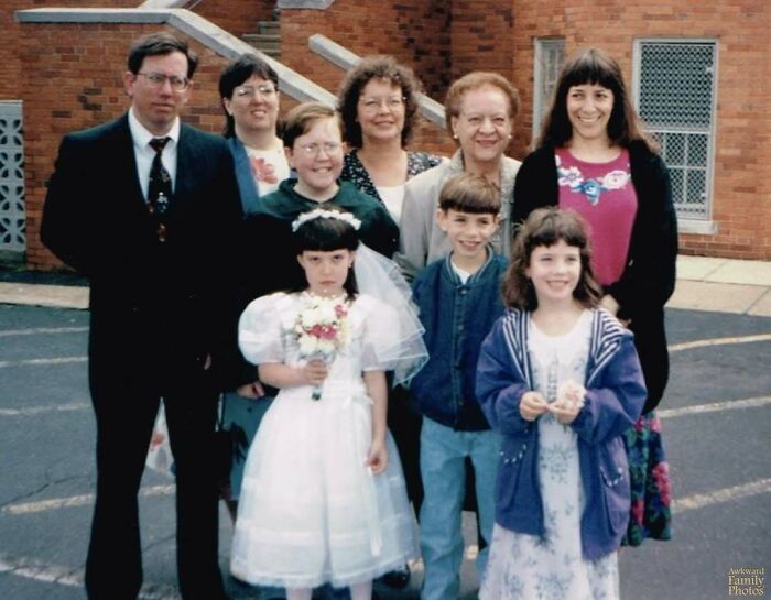 Awkward family photo with eight people outside a church, including a girl in a white dress holding flowers.