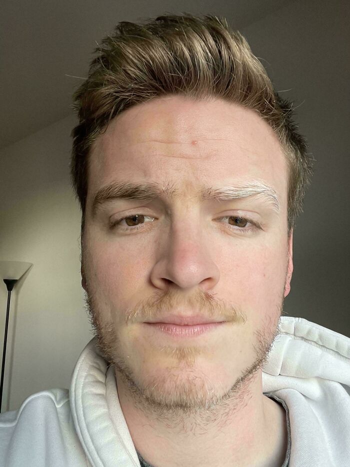 Close-up portrait of a young man with light hair and a slight beard, wearing a white hoodie.