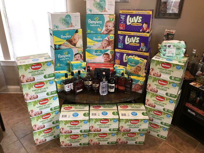 Stacks of diapers and bottles of alcohol create a funny pregnancy situation in a living room setting.